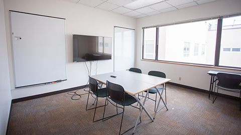 View into a room brightly lit with sunlight with two tables and chairs in front of a wall mounted screen and two whiteboards.