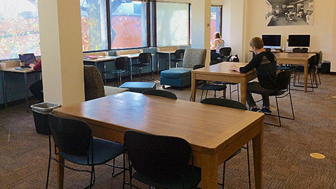 Students working at tables in a open area with windows to the side.