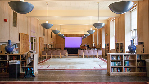 A large classroom with bookshelves on either side and a projector screen in front of rows of chairs.