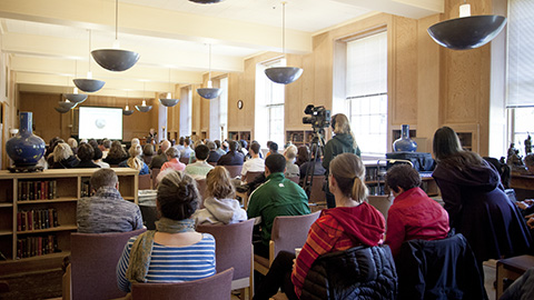 A classroom of people listening to a lecture in a room with large windows.