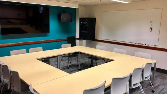 Classroom with wall mounted screen, two large speakers, and tables and chairs arranged in a square.