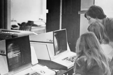 Three people sit at giant, old computers. 