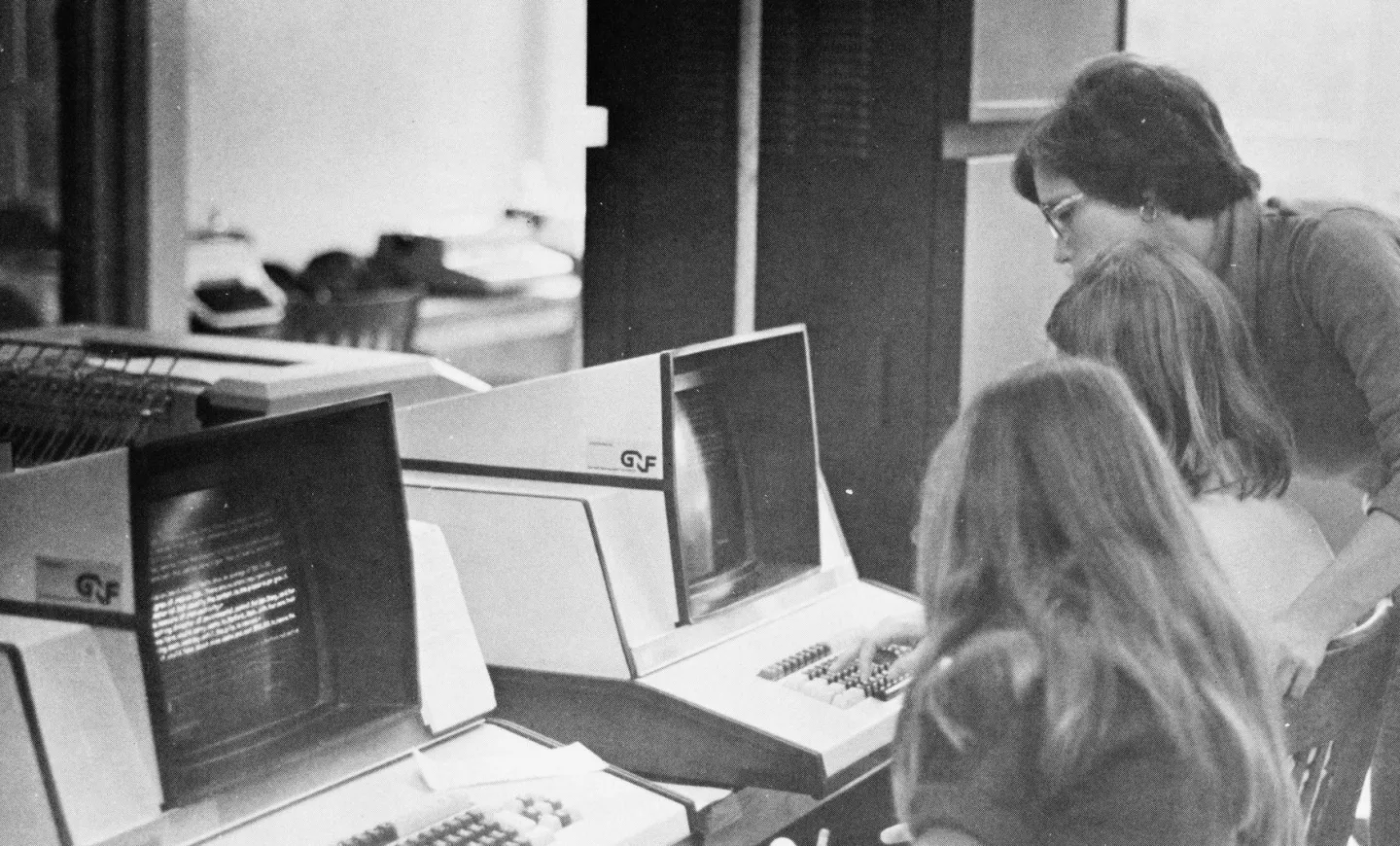 Three people sit at giant, old computers. 